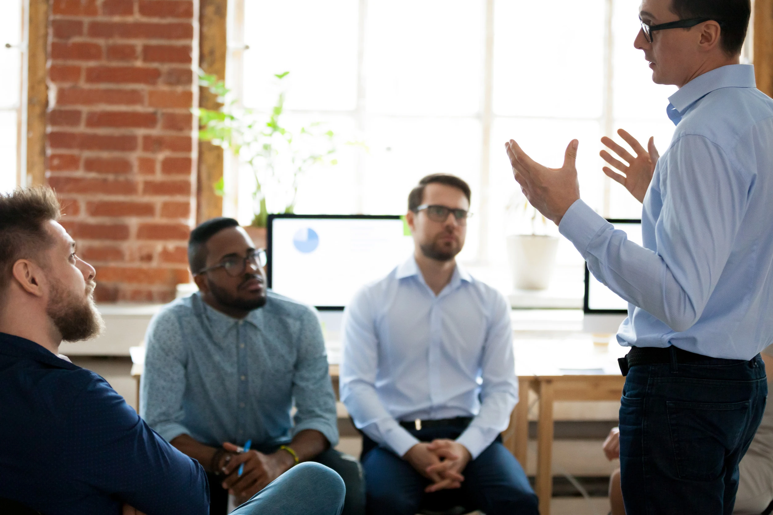 Confident male leader mentor coach speaking at diverse team meeting, presenter speaker manager explaining new business plan strategy to employees group during office briefing at corporate training