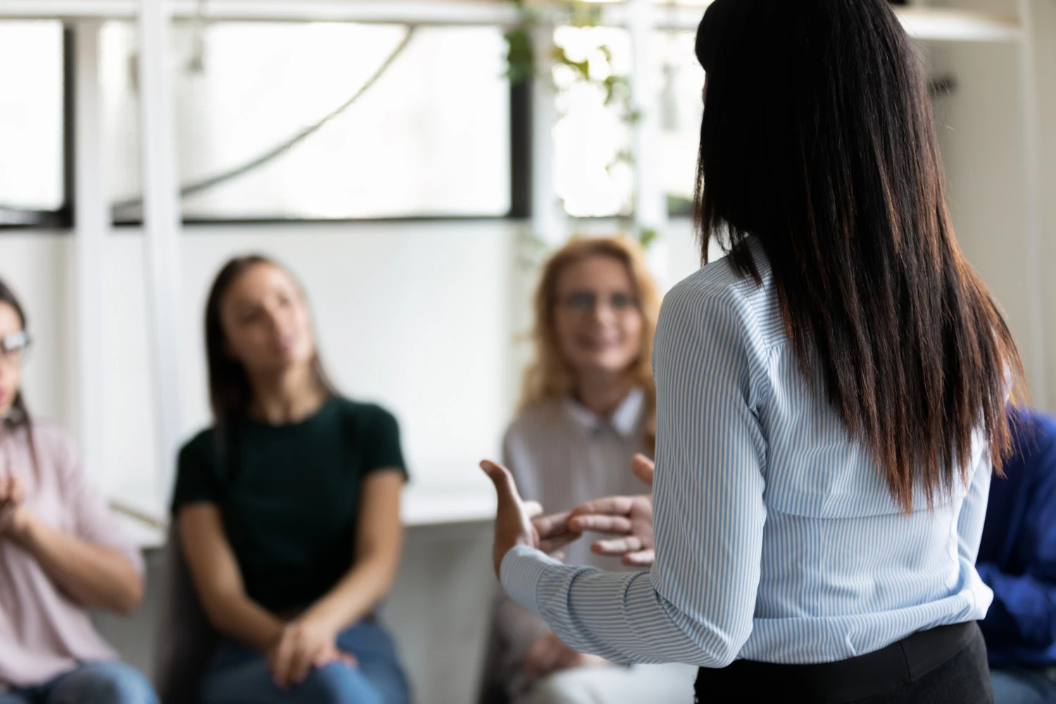 Rear back view female african ethnicity participant at group rehab session telling story share personal problem with associates or corporate staff gathered together listening business coach at seminar